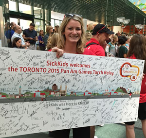 A woman holds a large banner covered in signatures. The banner reads 'SickKids welcomes the Toronto 2015 Pan Am Games Torch Relay" the 