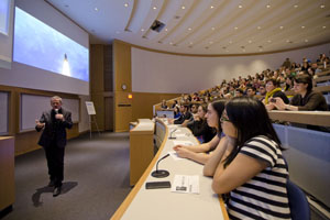 Man stands at the front of a lecture hall filled with people.