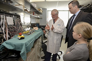 James Drake, wearing a white SickKids lab coat, explains a large piece of technical machinery to a man and child who observe attentively