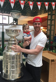 A man holds a child next to the stanley cup, a large silver trophy