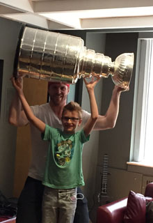 A boy holds the stanley cup, a large silver trophy, above his head, with the help of a man standing behind him