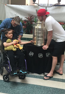 A child in a wheelchair touches the stanley cup, a large silver trophy, while talking to Bryan Bickell