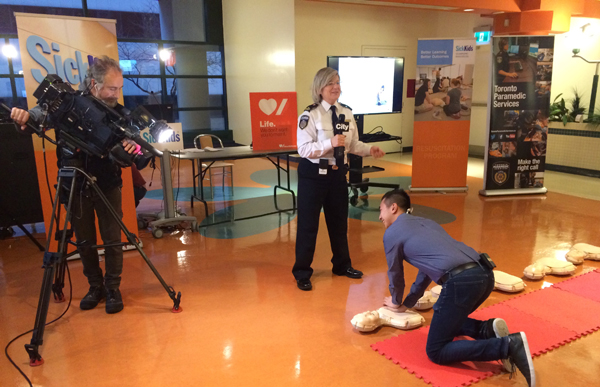 A camera man films an adult doing chest compressions on a CPR dummy while someone dressed in paramedic gear instructs through a microphone