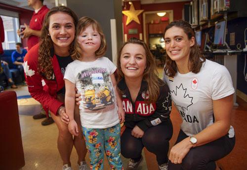 Three women wearing Team Canada clothing kneel and smile with a young girl