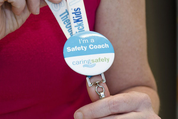 A button on a lanyard. The button reads "I'm a Safety Coach." with the Caring Safely logo