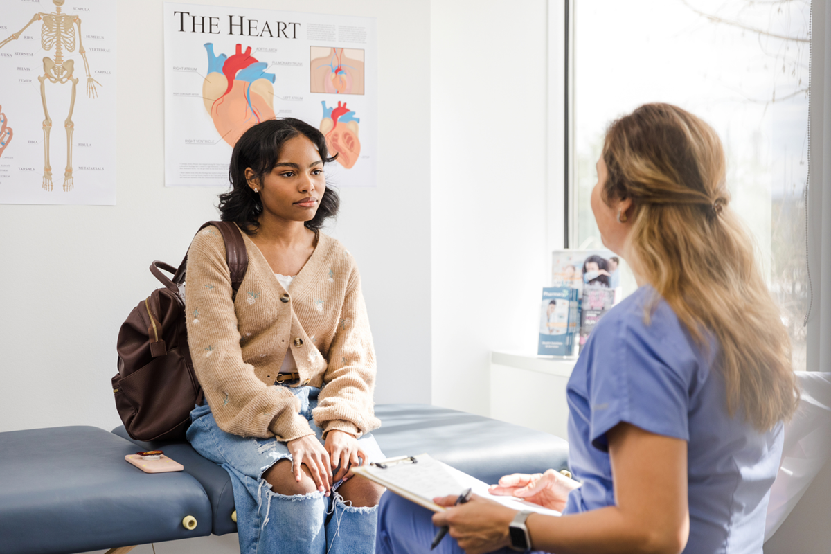 An adolescent sits in a medical room looking at a care provider.