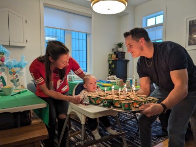 Erica stands behind Jeff, sitting in a high chair, while Brett holds treats with candles on them. 