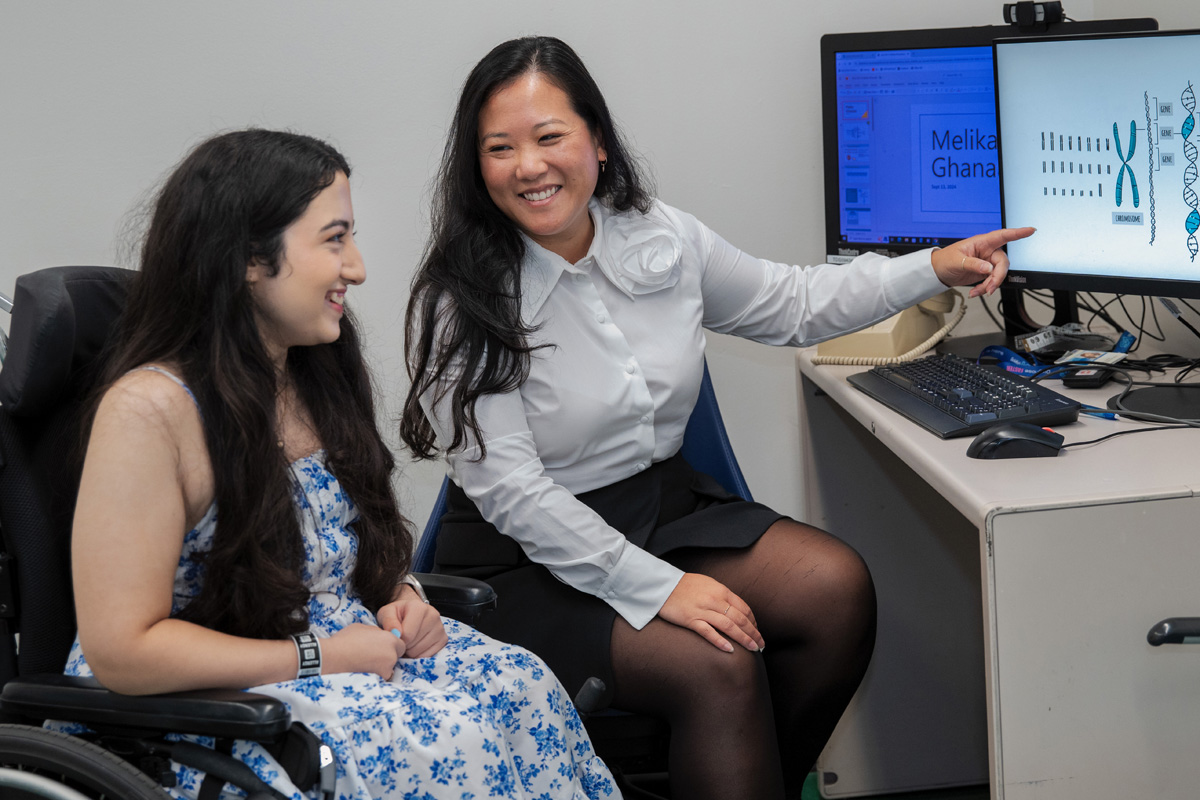 Melika and Anna are smiling and seated by a computer. Anna points to an illustration of a chromosome on the computer screen.