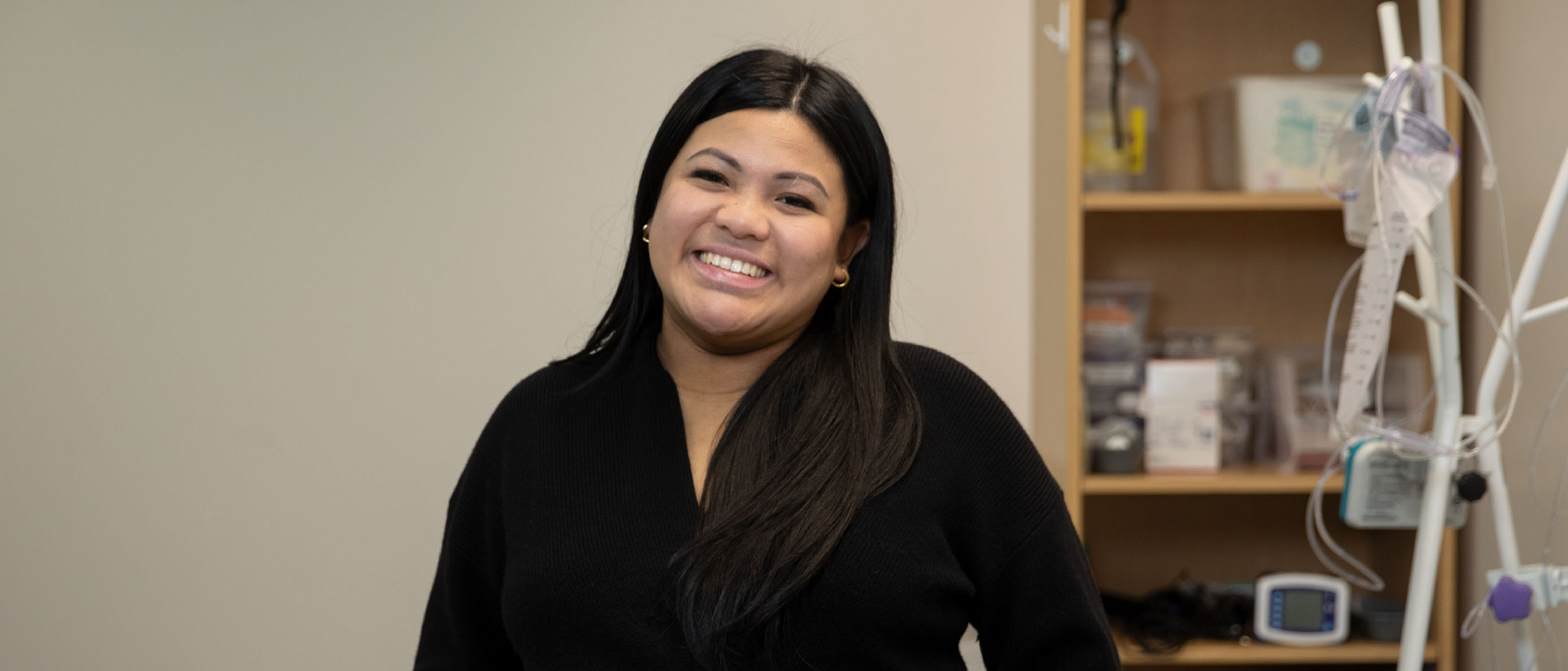 Bianca Dolor smiling for a photo. In the background, there is an IV stand and a shelf containing various medical supplies.