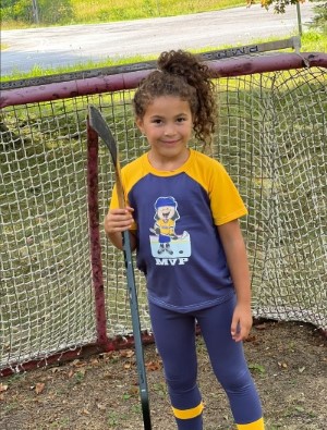 A girl smiling and holding a hockey stick and standing outside in front of a hockey net. Her T-shirt has a character in hockey gear with the text "MVP" underneath,