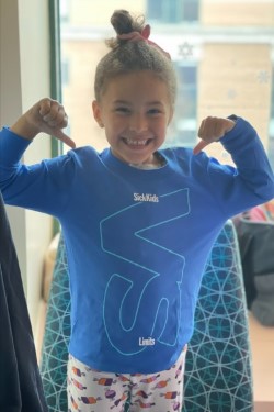 A young girl wear a blue SickKids VS shirt. She has her arms up and thumbs pointing toward her shirt.