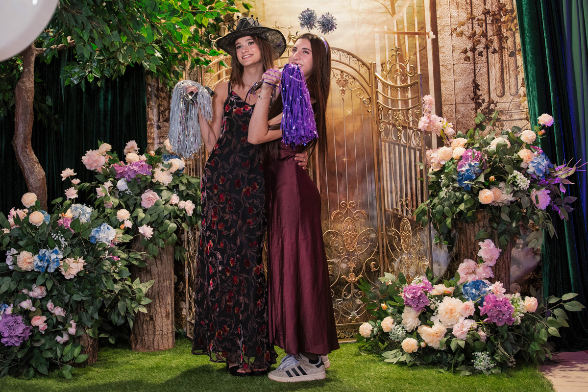 Two SickKids' prom attendees in dresses. They pose for a photo together in front of an enchanted forest-themed backdrop, with flower bushes, trees and a golden gate.