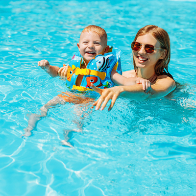 A child and mother in a swimming pool and smiling at the camera. The child is wearing a life jacket.