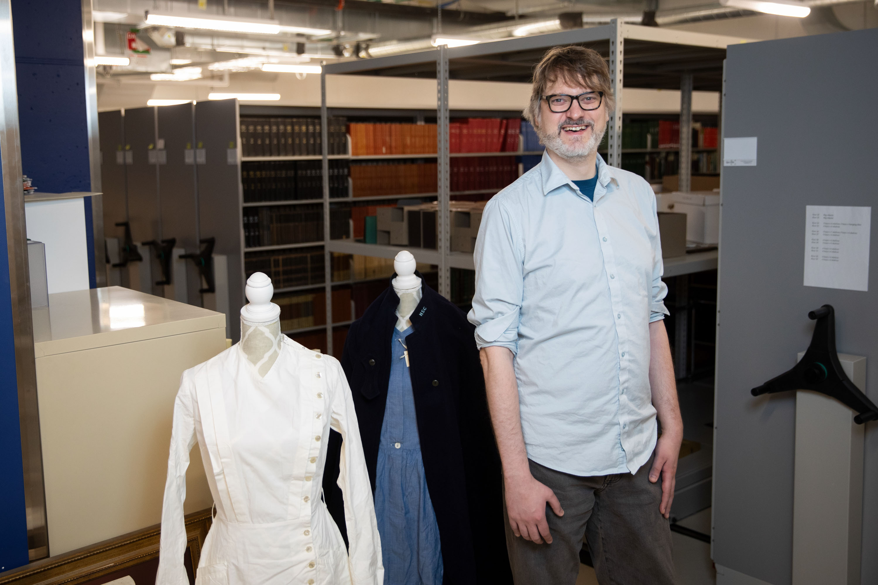 Man with glasses and light blue shirt standing in archive storage area with historical clothing displayed on mannequins nearby.