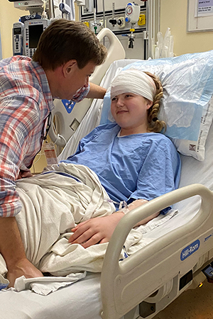 A young women lies in a hospital bed, her head bandaged after surgery, smiling at her father.