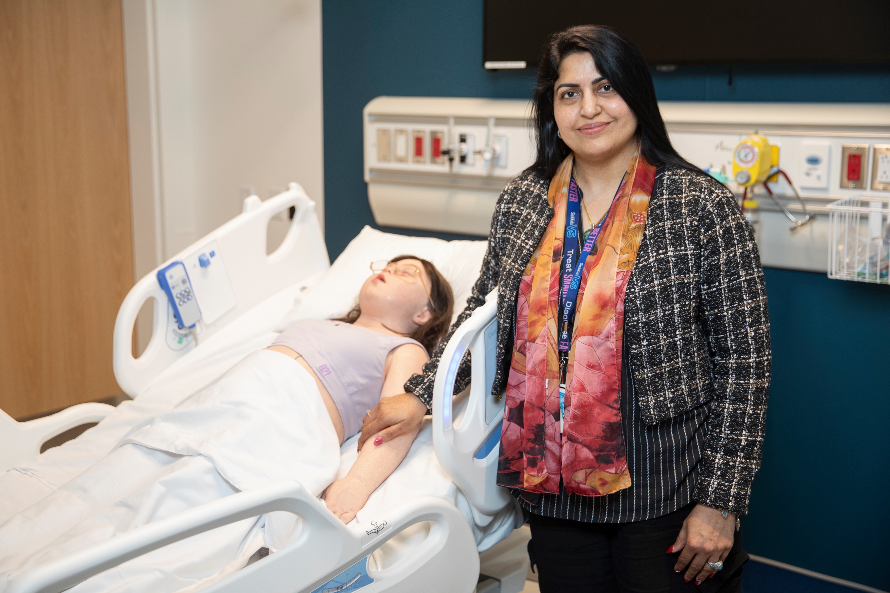 In the simulation centre, Dr. Jabeen Fayyaz, wearing a black plaid blazer and pink and orange scarf, stands next to a hospital bed where a manikin is lying.