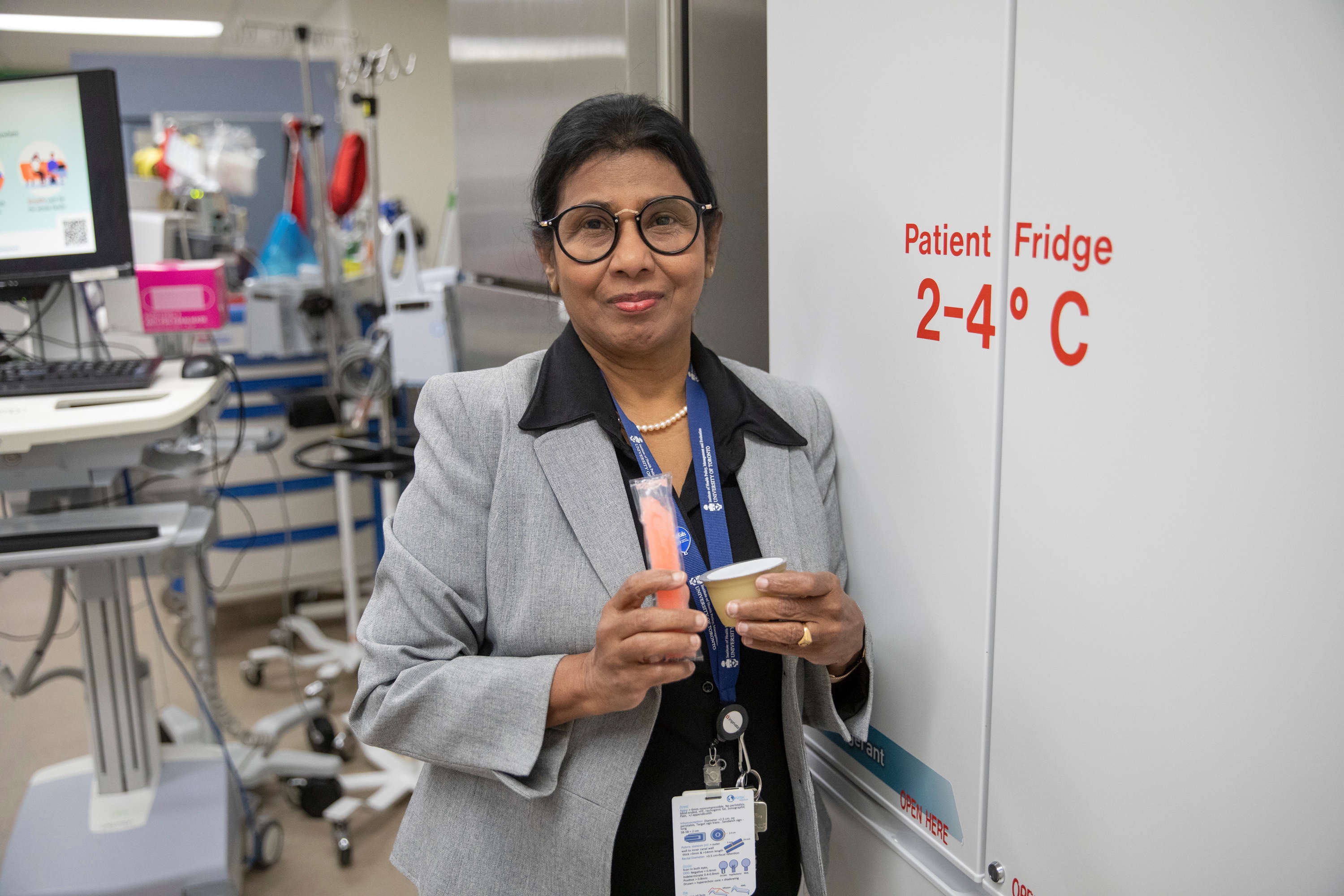 Dr. Savithiri Ratnapalan, wearing a black collared shirt and grey blazer, stands in front of a fridge holding an orange popsicle and apple sauce. 