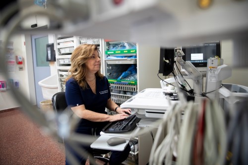 Dr. Suzan Schneeweiss is framed by an electrical cord in the foreground and she works on a computer in the emergency department in the background.