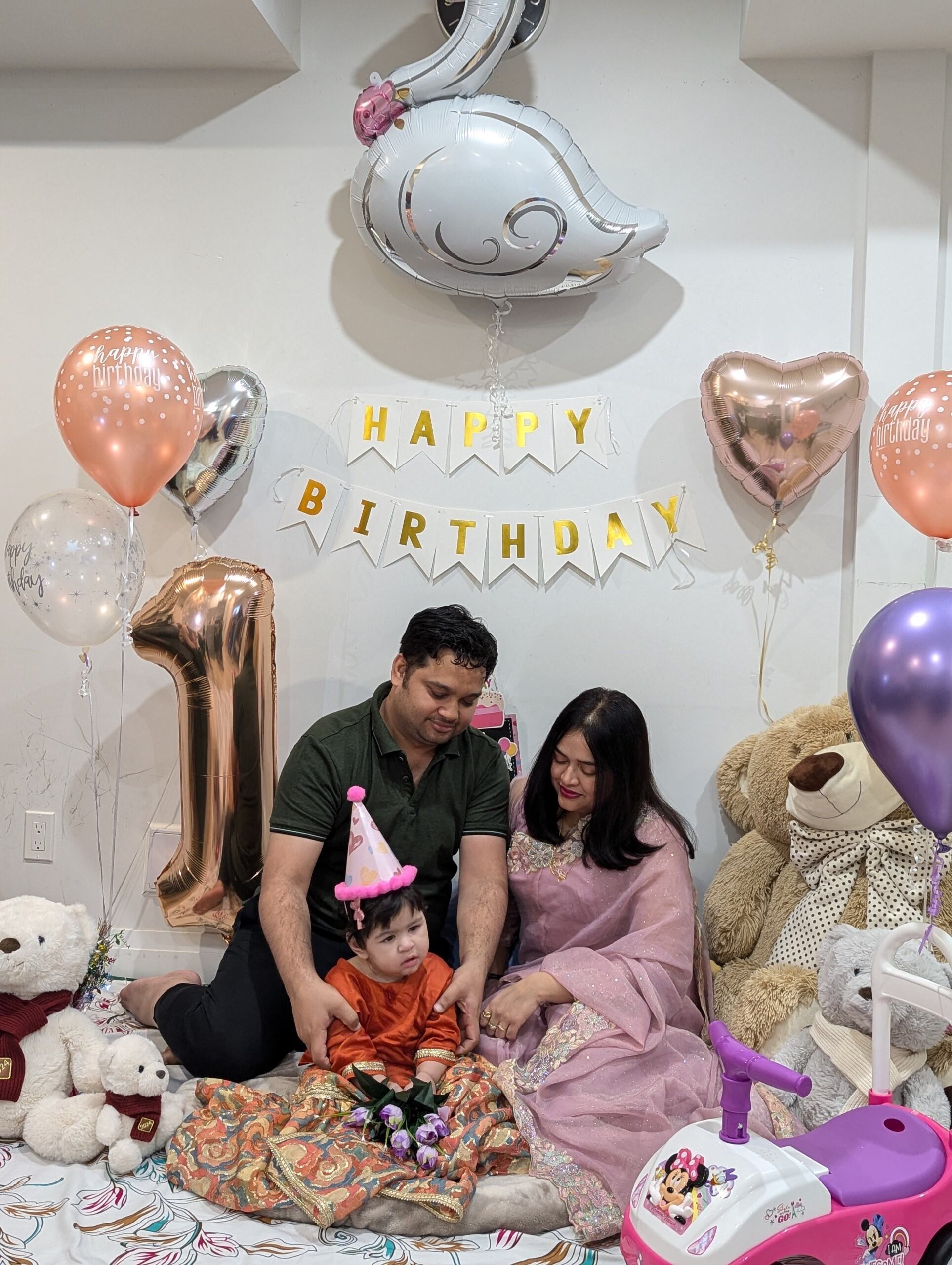 Family celebrating child's first birthday with rose gold balloons, "Happy Birthday" banner, and toddler in party hat surrounded by toys.