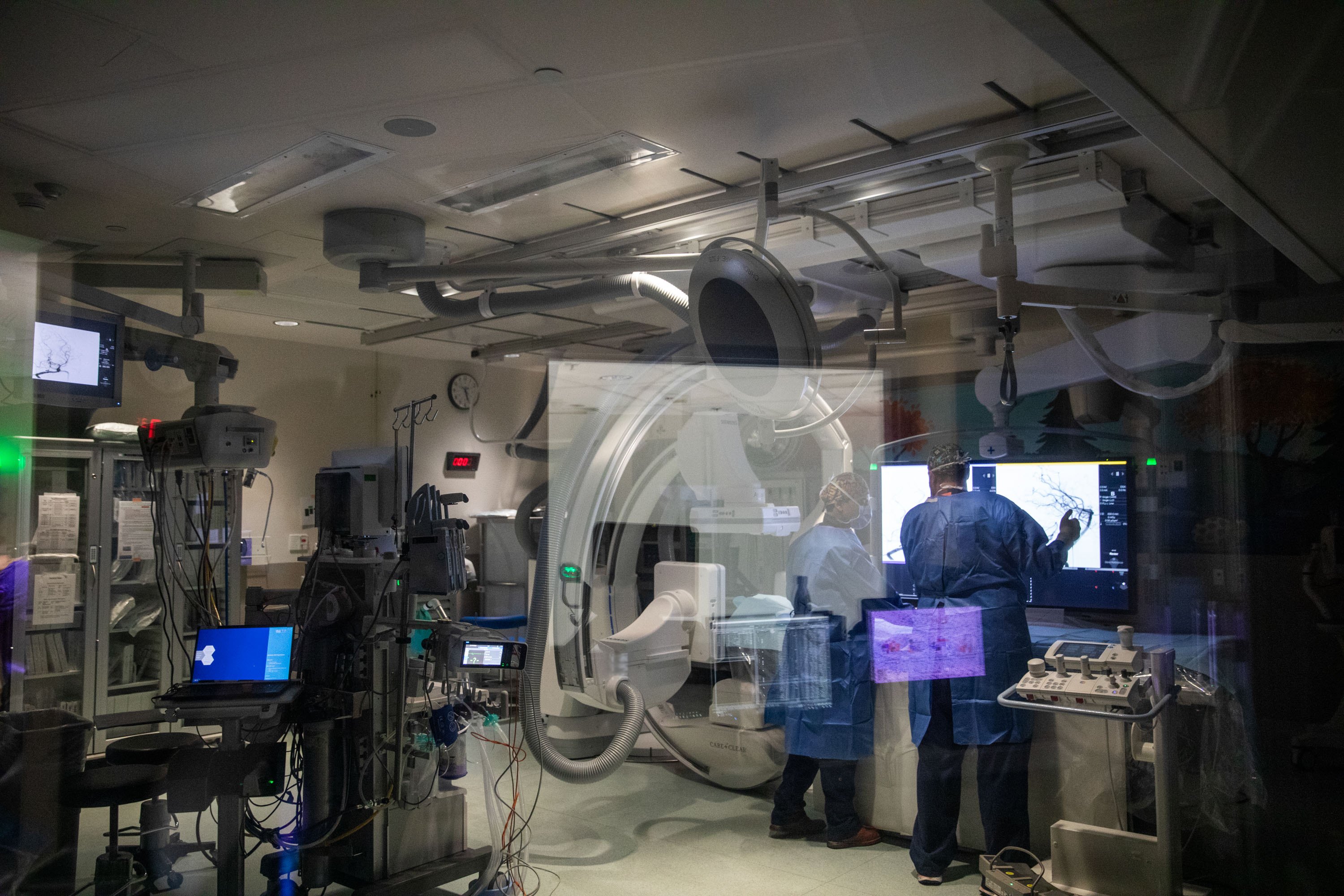 Advanced cardiac catheterization laboratory with C-arm fluoroscopy equipment and medical staff performing interventional procedure with multiple monitoring screens.