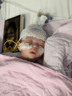 Baby in hospital bed wearing knit cap and nasal tube, resting on pink bedding.