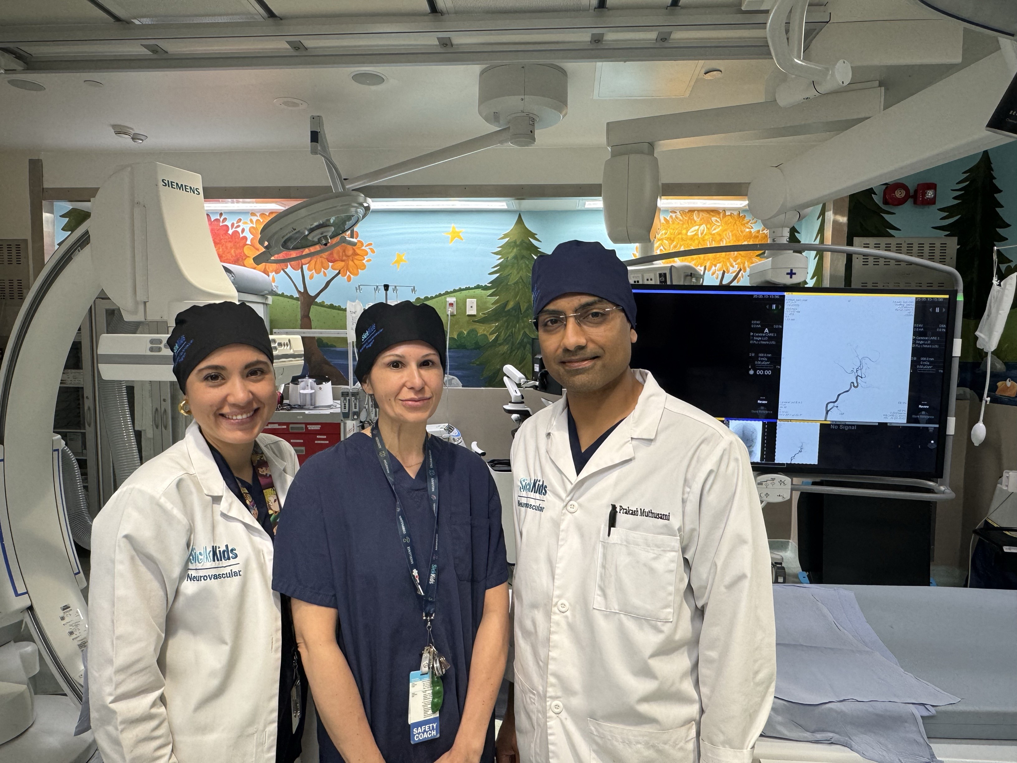 Three medical professionals posing in pediatric catheterization laboratory with colorful nature-themed wall murals and Siemens imaging equipment.
