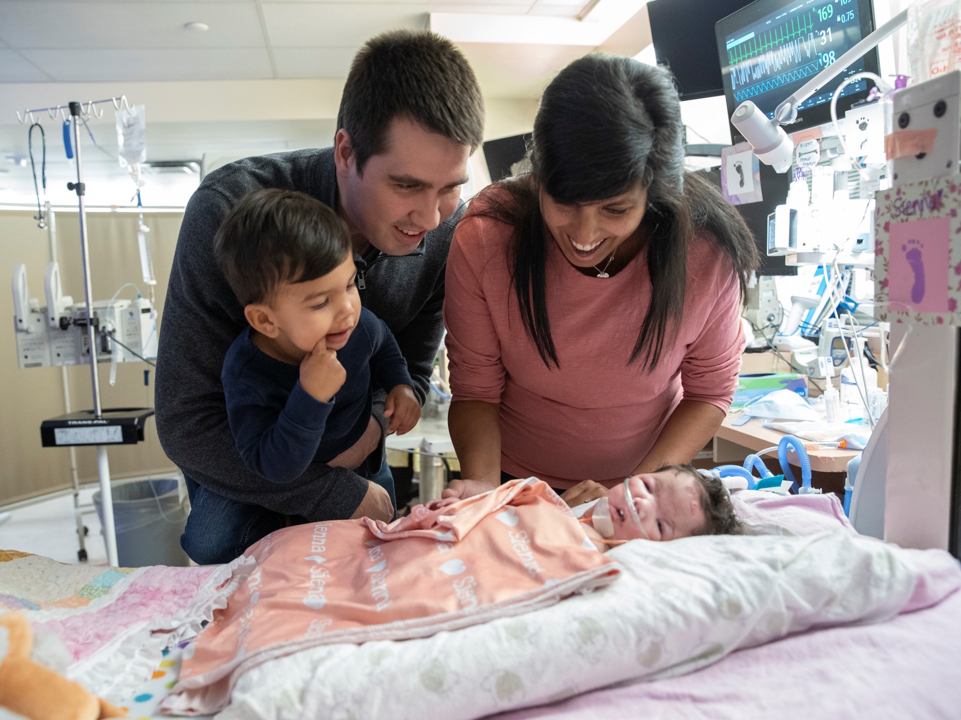 Sienna is wrapped in a peach coloured blanket, lying down in the NICU. Brittany, Tyler and Camden all stand at her side and look at her, smiling.
