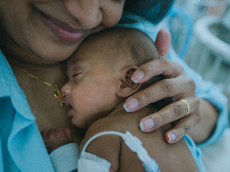 Person smiling while holding baby with medical tubes on arm, resting on their shoulder.