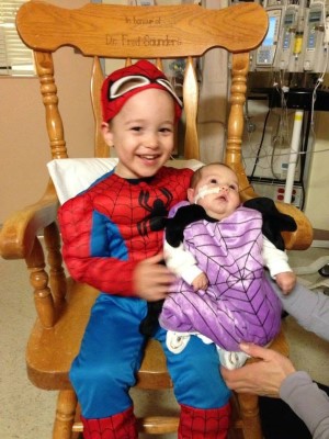 On a wooden chair, Leah's son is dressed as Spiderman, and holds Leah, who is wearing a purple spider costume.