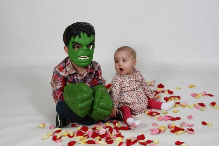 Leah looks at her brother, who is wearing a green superhero costume. The pair is sitting on a white background with red petals on the ground.