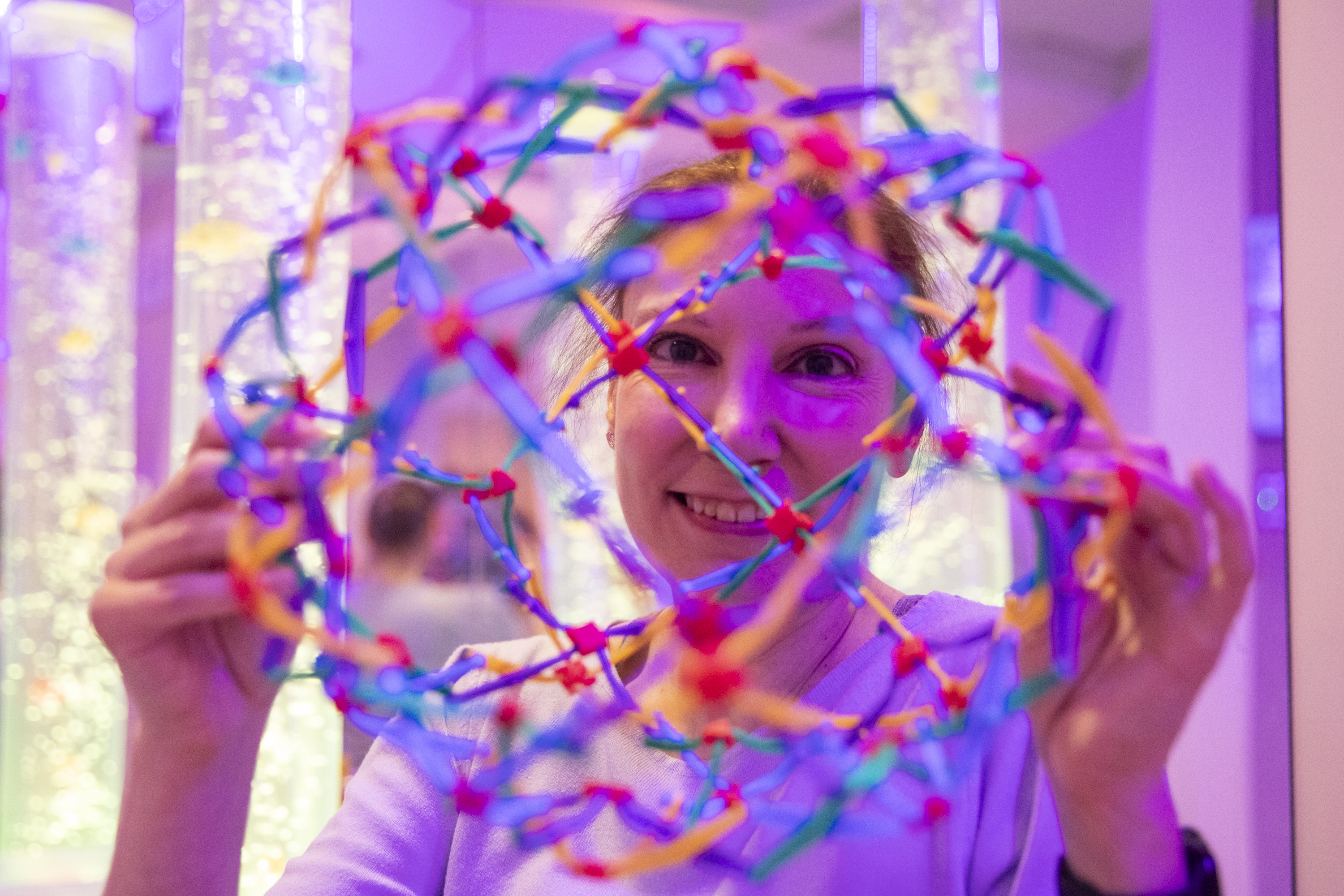 Natalie holds a spherical, colourful plastic toy that expands and compresses, in front of her face. There is a pink-purple light in the background.