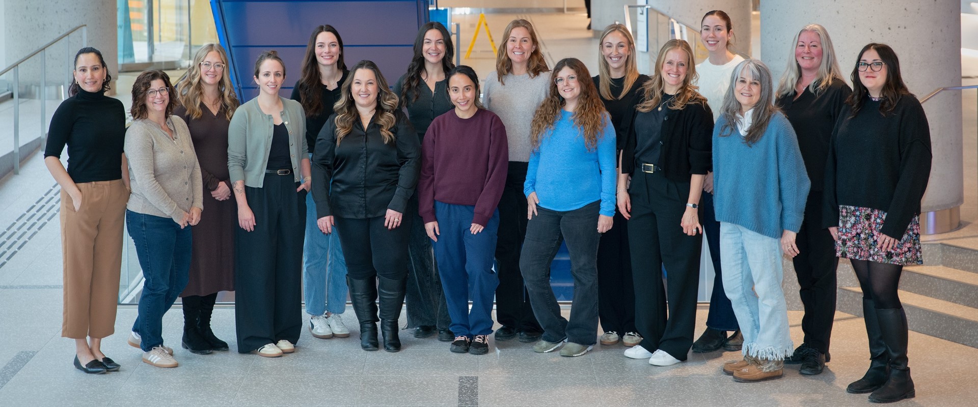 A group of people stands together indoors for a team photo. They are arranged in two rows in a bright open space with stairs in the background.