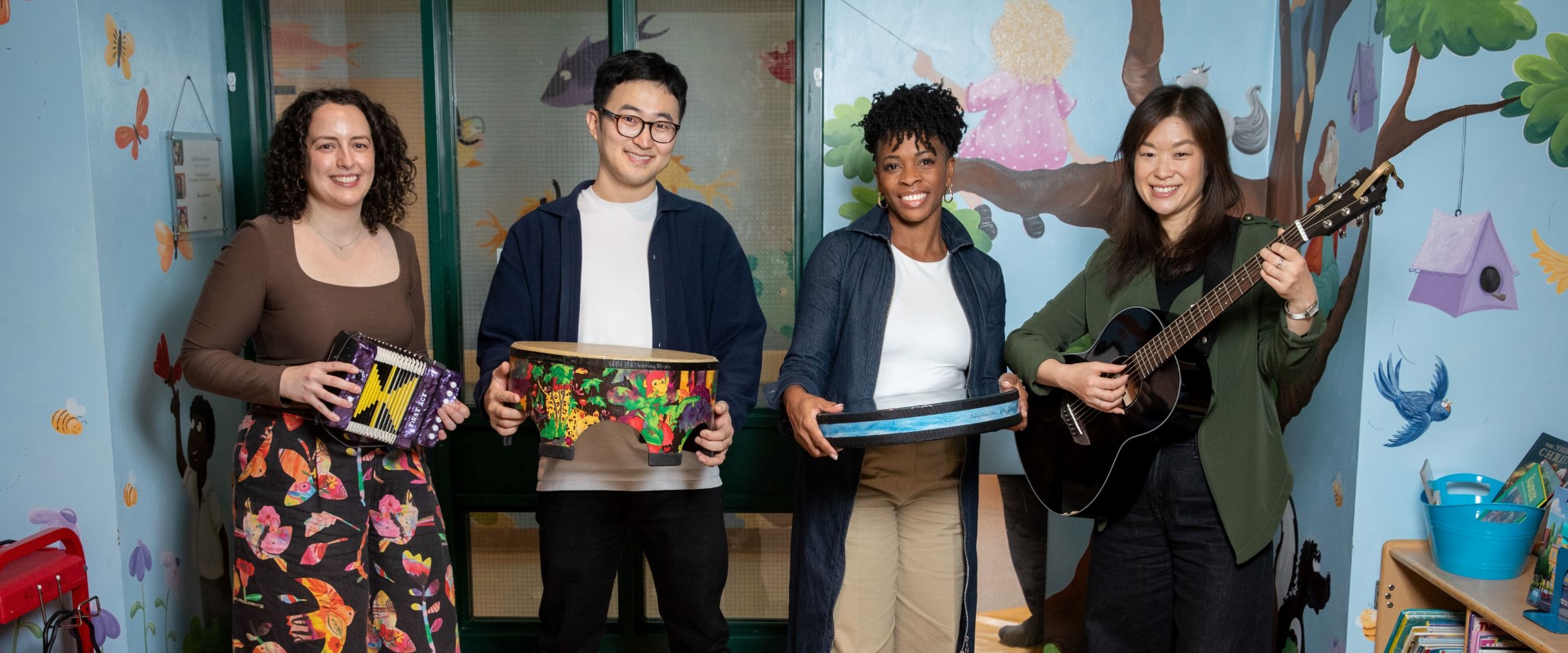 Four people standing on colorful mats in a children’s playroom holding musical instruments, including a guitar and drums.