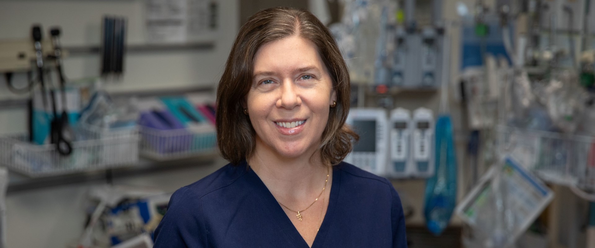 A person wearing navy medical scrubs standing in a hospital room with medical equipment in the background.