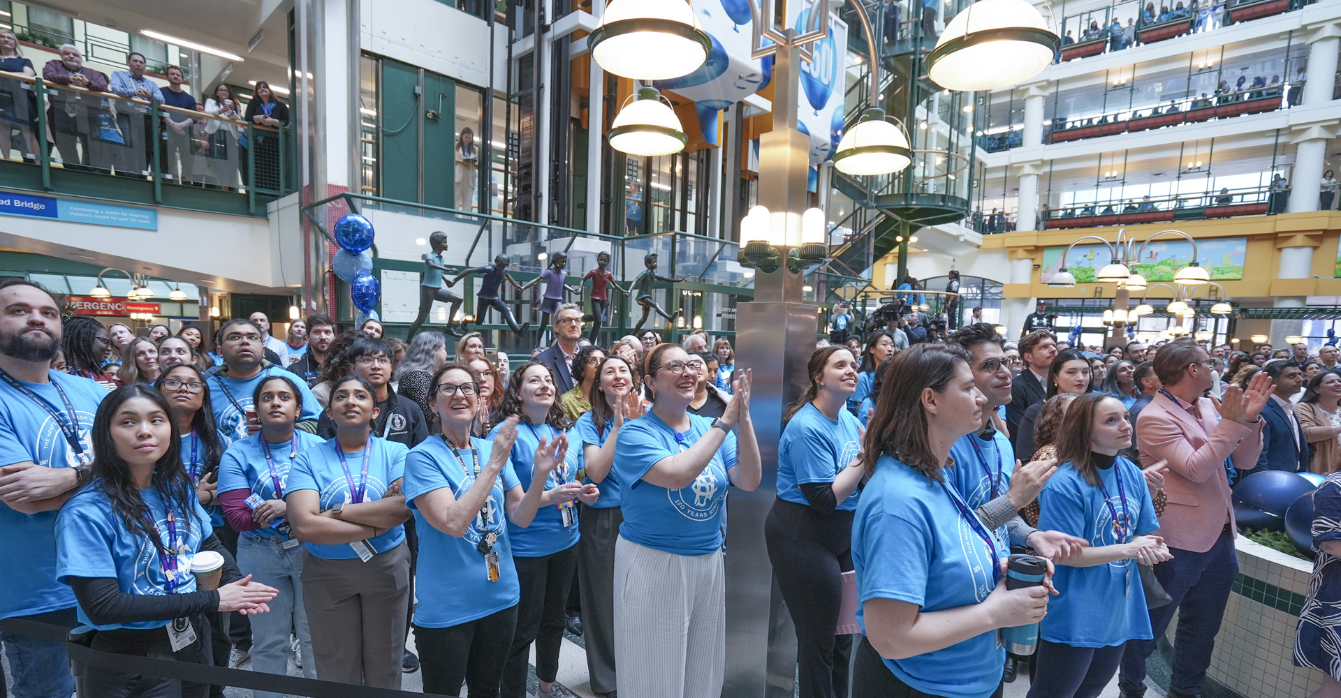 A crowd of people dressed in blue commemorative SickKids 150 t-shirts applauding in the Atrium.
