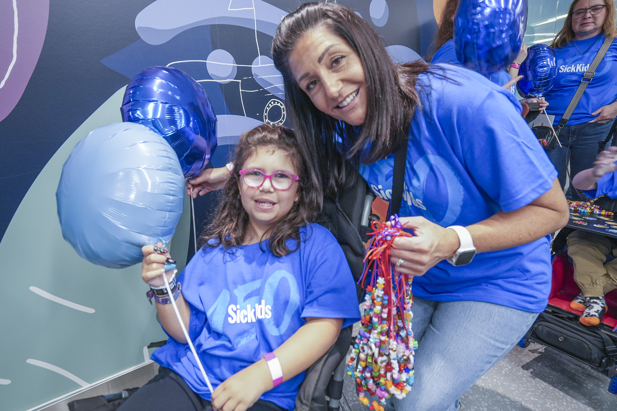 Morgan and Chloe posing for a photo. Both are dressed in blue SickKids 150 t-shirts and holding blue balloons. Morgan is carrying Bravery Beads in her other hand.