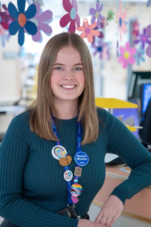 Melissa Darby stands smiling in front of a clinic desk with flower decorations hanging above her head. There are many bright pins on her SickKids lanyard, including one that reads Social worker.
