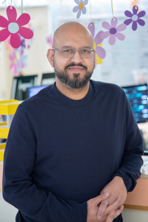 Dr. Aamir Jeewa stands smiling in front of a clinic desk with flower decorations hanging above his head.