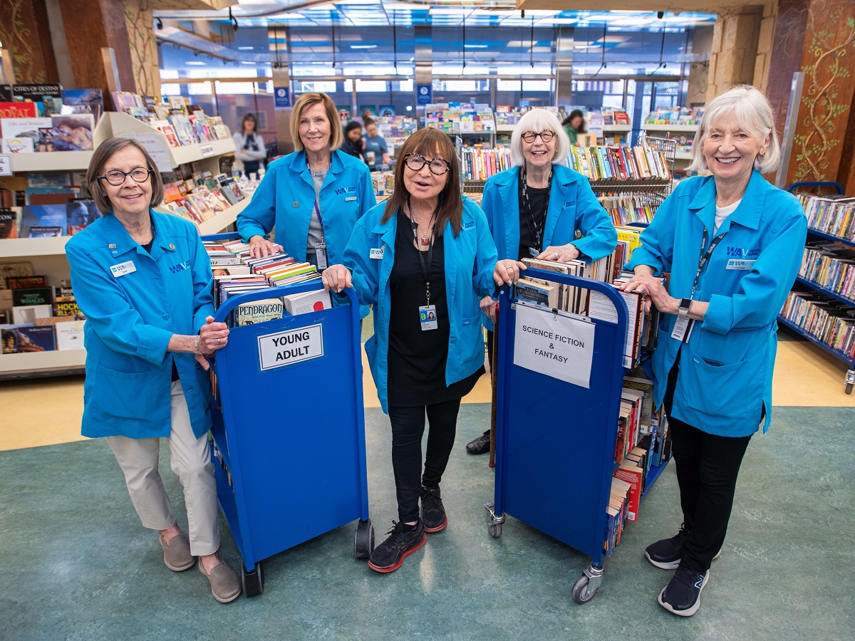 Five women in blue jackets stand with carts filled with books.