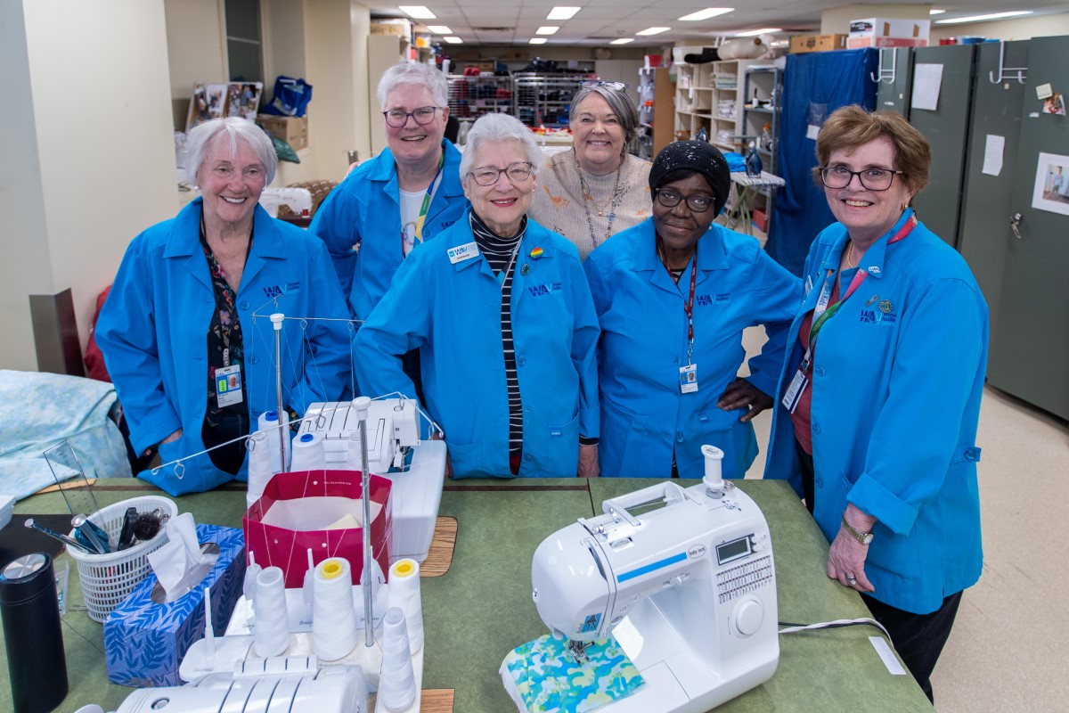 Five women in blue jackets pose together at a sewing table.