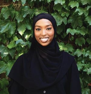 Riham Farah stands smiling in front of green foliage and is shown from the shoulders up. She is wearing a black dress and black hijab.