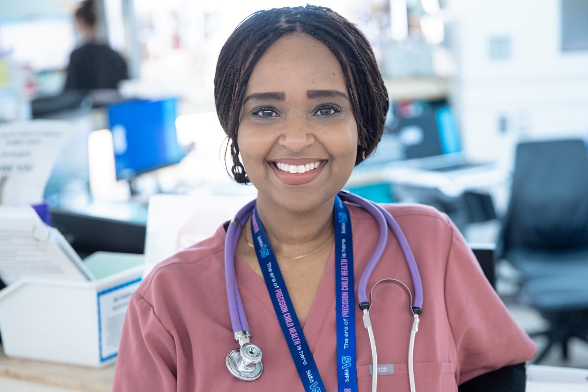 A healthcare worker wearing pink scrubs and a stethoscope stands at a hospital desk, with medical supplies and staff visible in the background.
