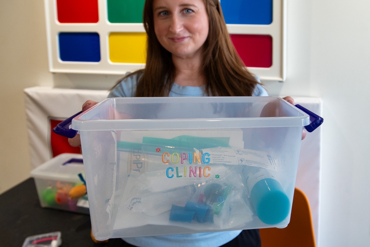Brianna holds a plastic box of supplies with colourful stickers on the front that read "Coping Clinic."