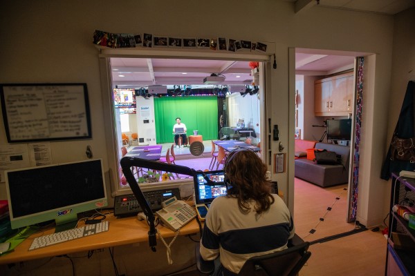 Emma runs the tech booth in a room attached to the studio while, in the background, Caleb leads bingo in front of a green screen.