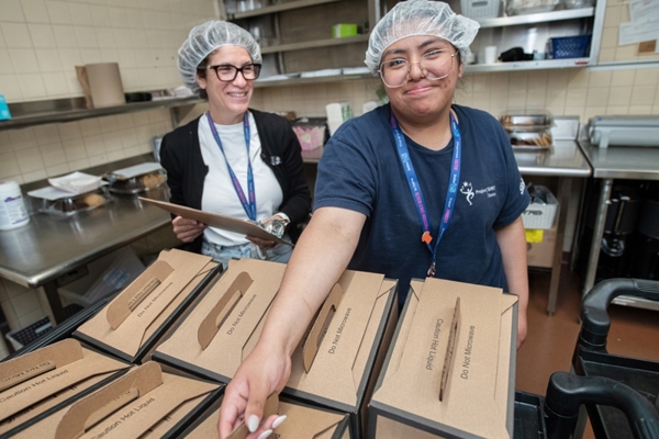 Woman reaches forward to hand an item across a table filled with packaged meal trays while another staff member stands nearby.
