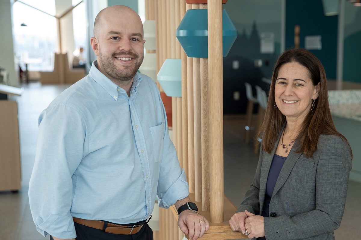 wo adults stand together in a modern, brightly lit room with wooden posts and colorful hanging shapes.