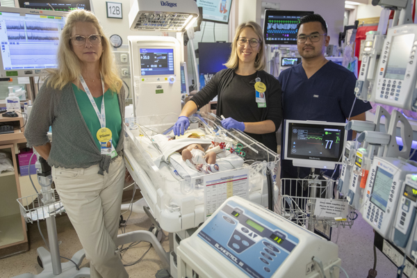 Three nurses pose for the camera and stand around an infant patient sleeping in a NICU bed.