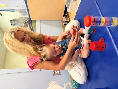 A nurse sits behind a toddler on a blue mat playing with toy blocks.