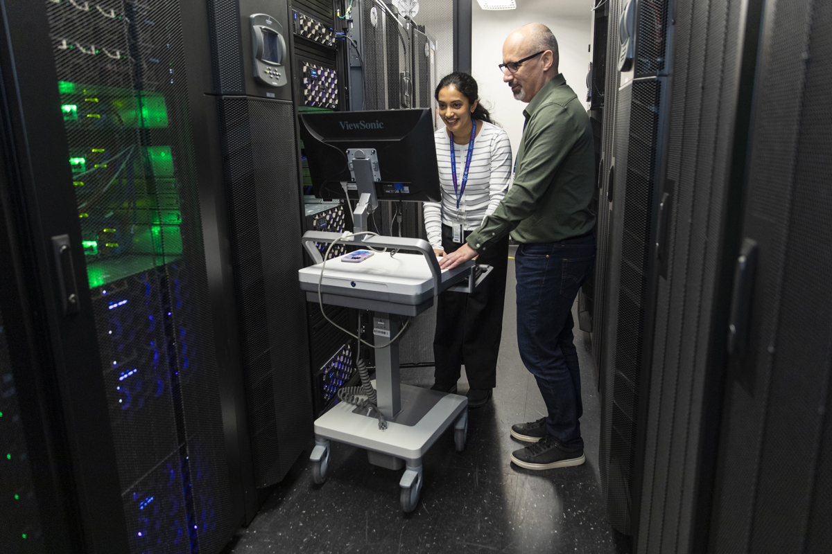 Two technicians stand in a data centre aisle, working at a mobile workstation surrounded by tall server racks with glowing indicator lights
