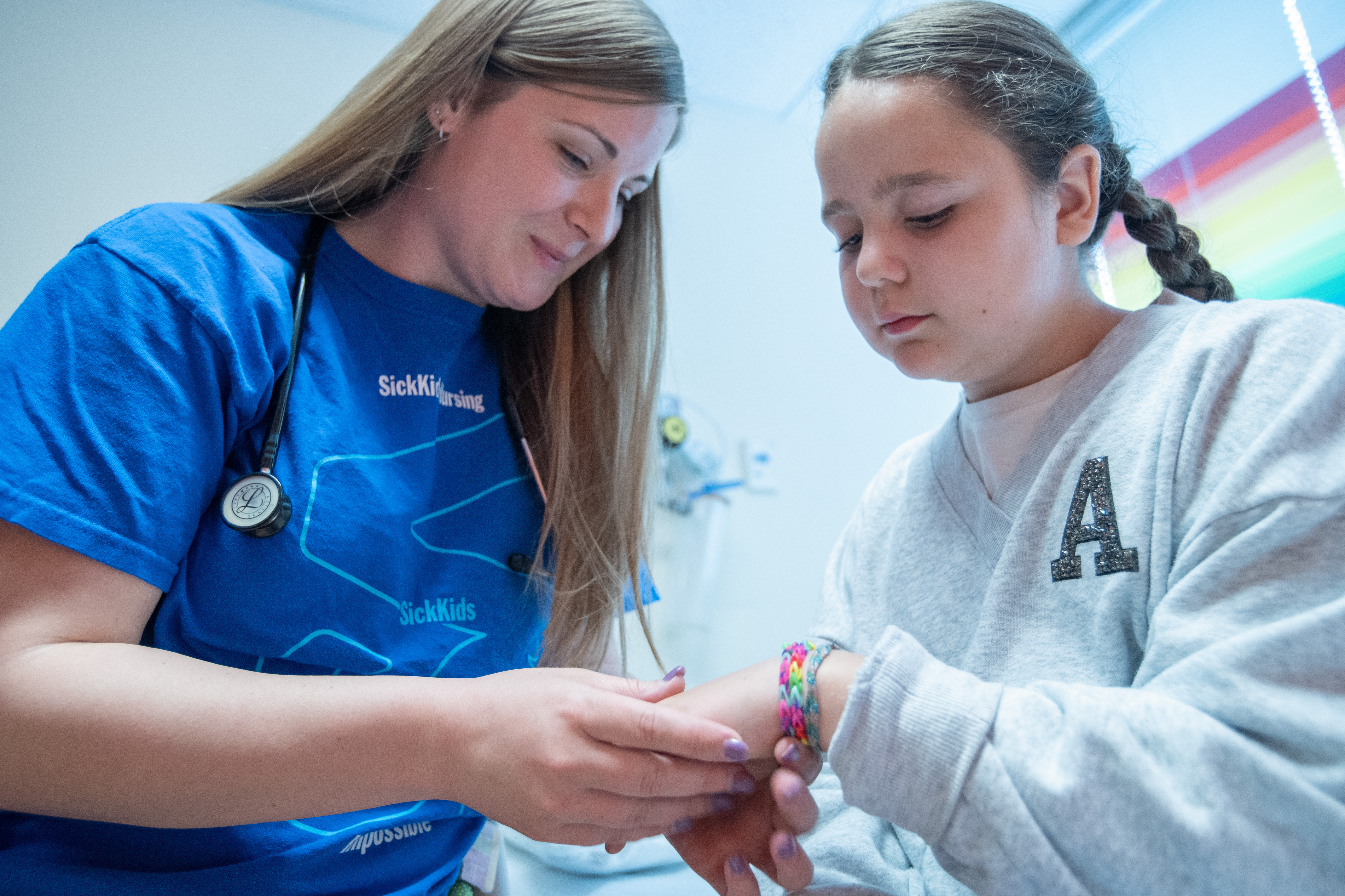 Healthcare worker in blue scrubs with stethoscope gently holding hands with a young patient in gray sweatshirt, providing comfort and care in a bright medical setting.